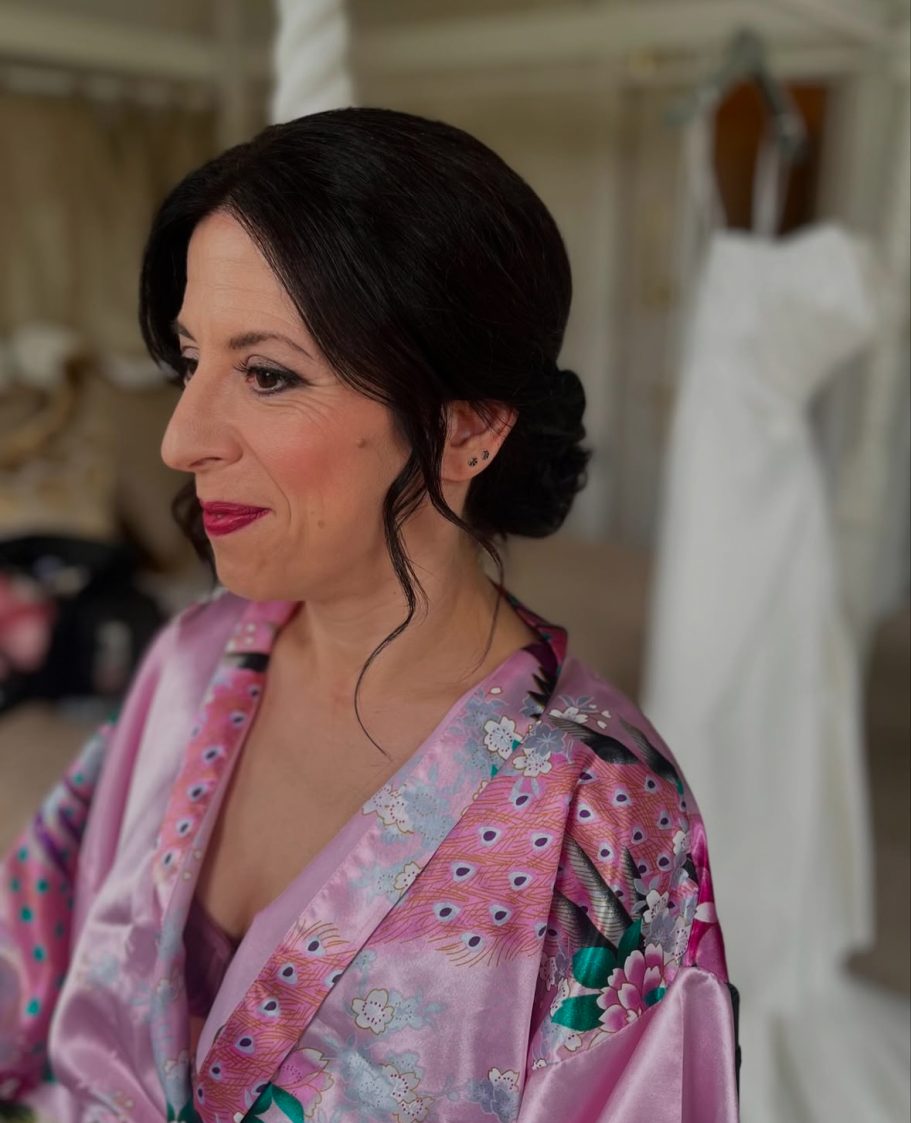 Woman in a pink robe with dark hair styled in a bun, looking thoughtfully to the side.Older bride, low bun, dark lip, smoky eye makeup